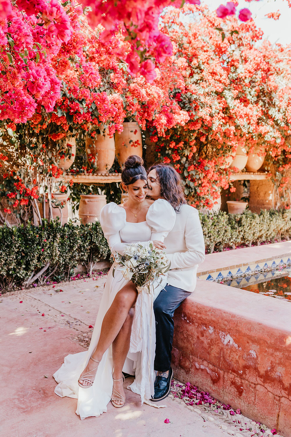 bride and groom sat on a wall surrounded by terracotta pots and bougainvillea flowers at Beldi Country Club in Marrakech