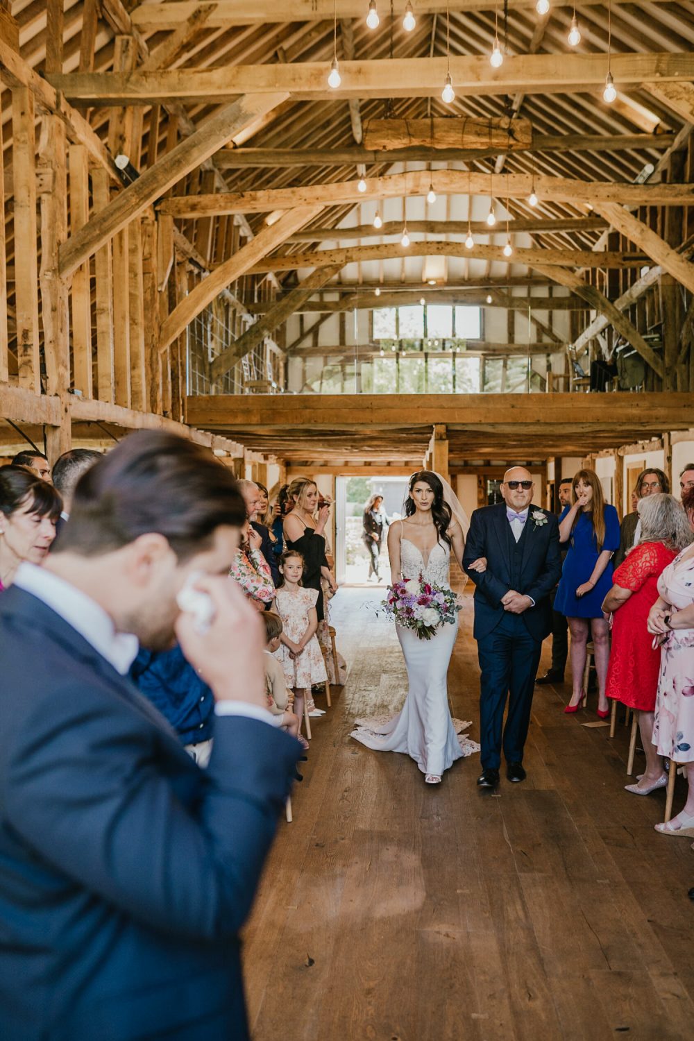 bride and her father walking down the aisle in the barn at blake hall in essex as groom cries