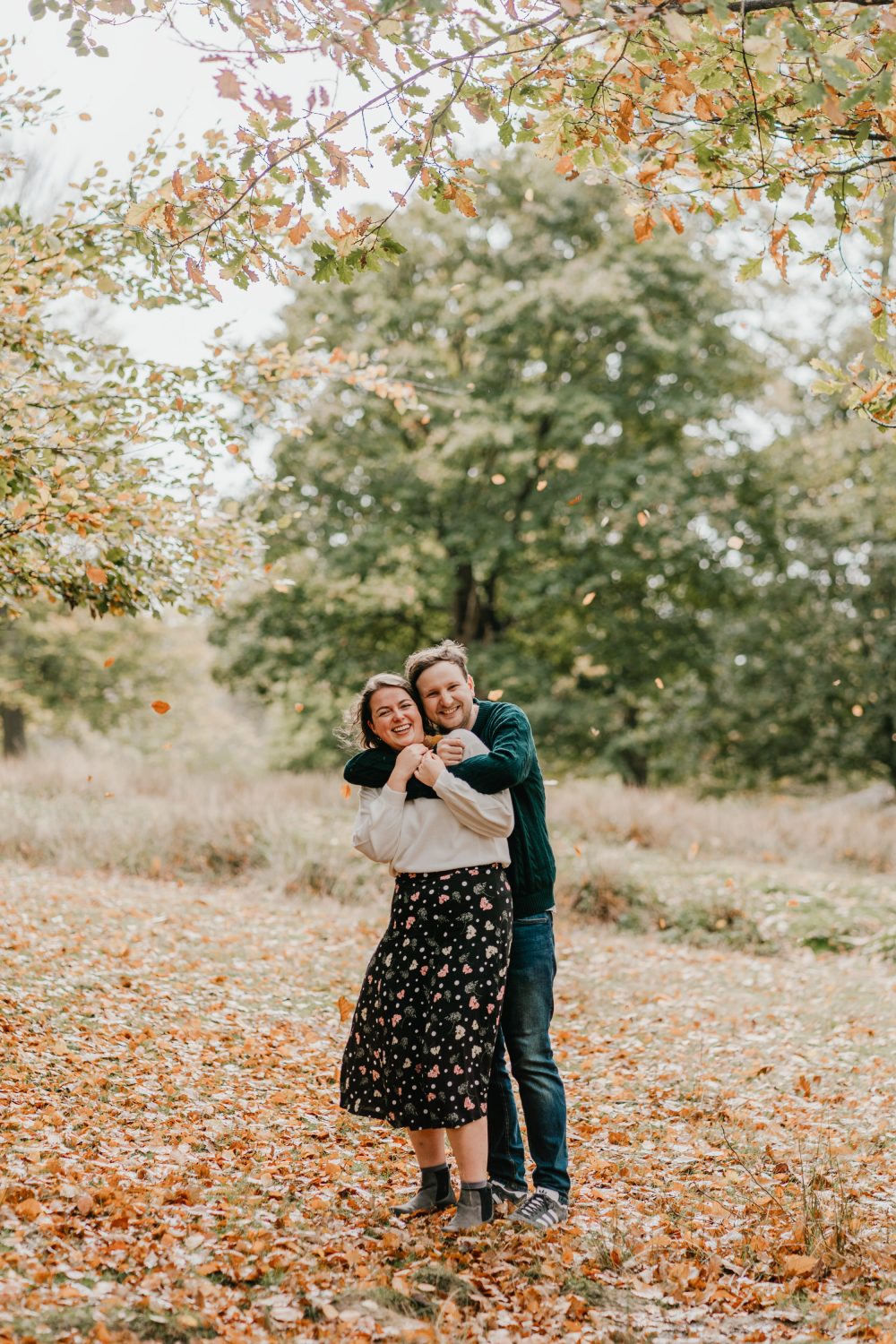 couple cuddle in the woods and laugh as leaves fall around them
