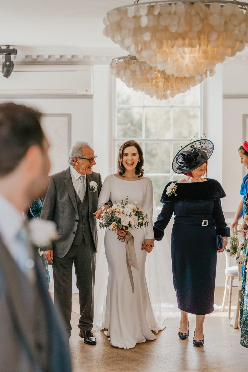 bride walks down the aisle with both her parents and smiles as groom turns to see her