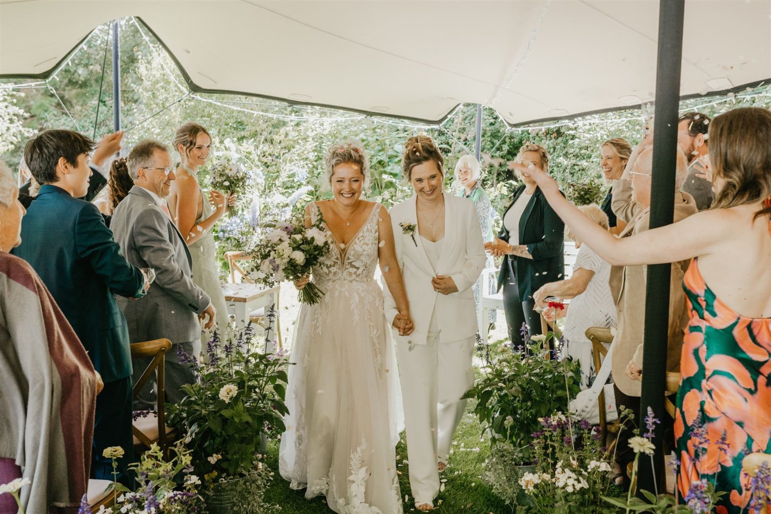 brides smile whilst their wedding guests throw confetti as they walk back up the aisle of garden wedding ceremony