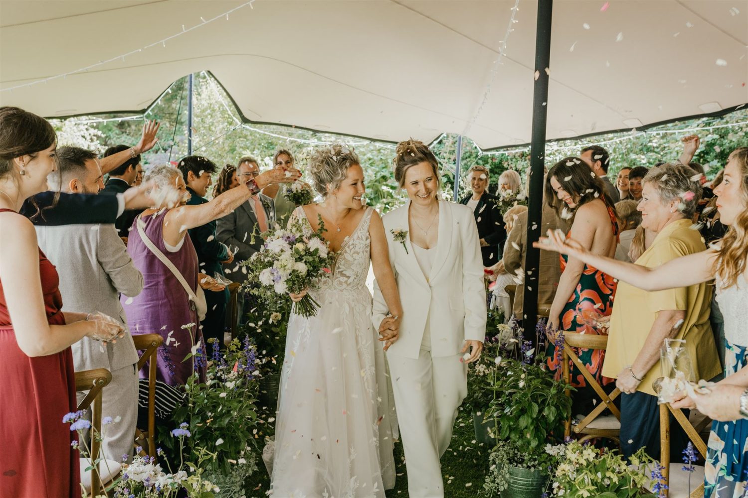brides smile whilst their wedding guests throw confetti as they walk back up the aisle of garden wedding ceremony