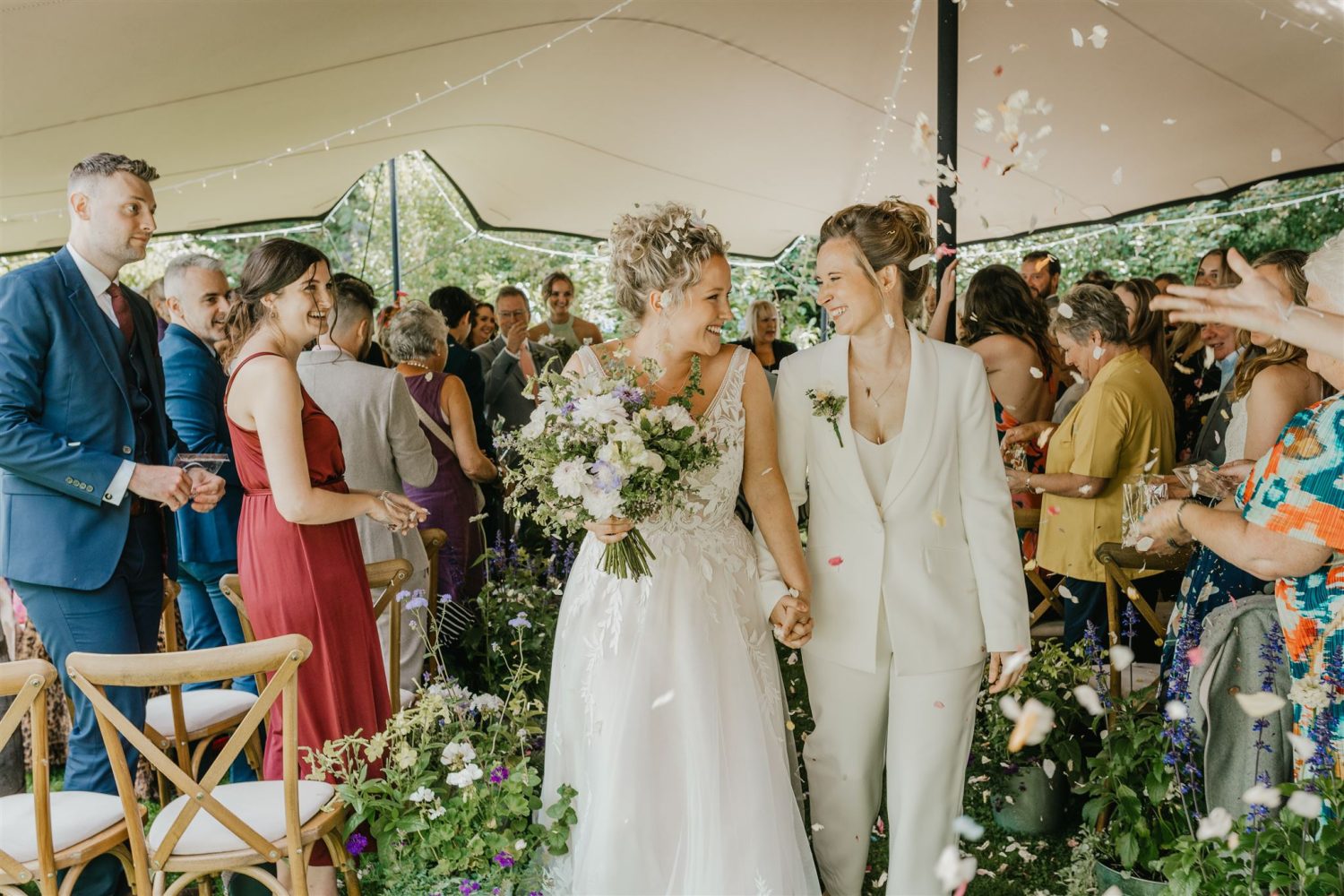 brides smile at each other whilst their wedding guests throw confetti as they walk back up the aisle of garden wedding ceremony