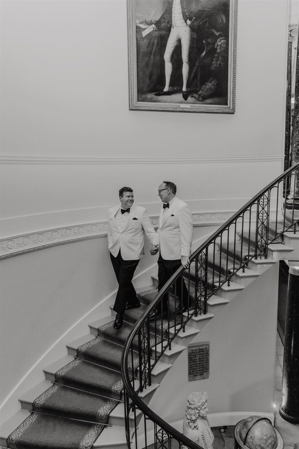 black and white photo of grooms descending the staircase at trinity house in london with white tux jackets