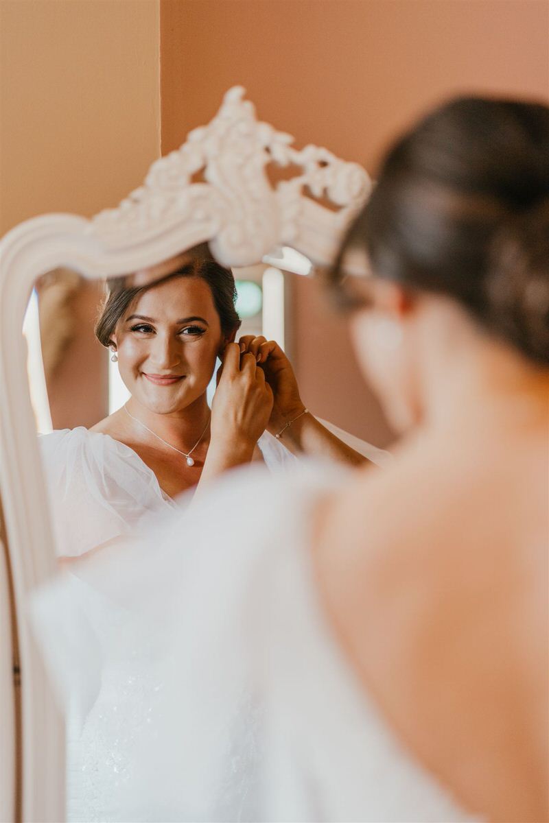 bride putting on her earrings whilst smiling in the mirror